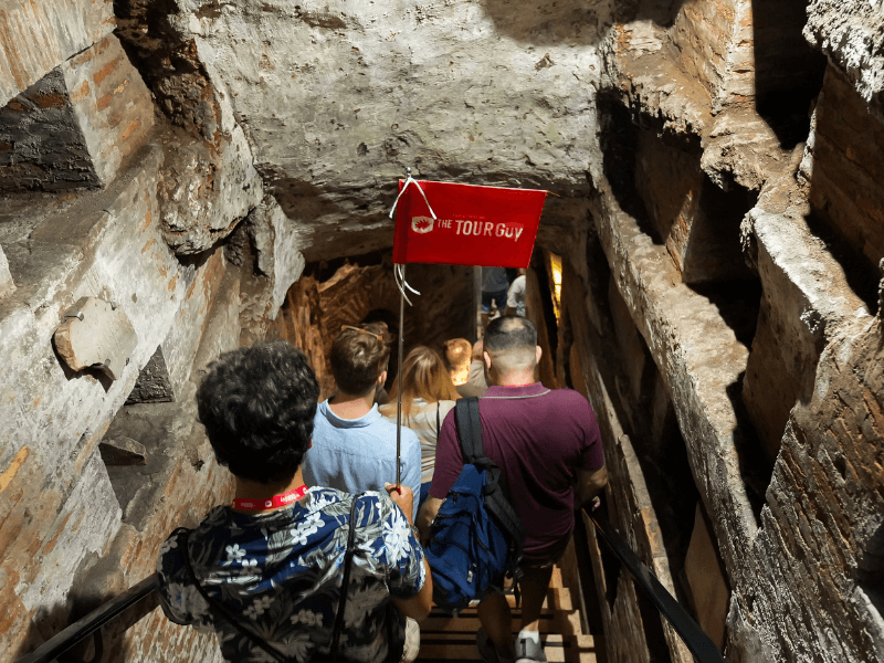 A group of tourists going down the stairs of a catacombs in Rome with their guide from The Tour Guy.