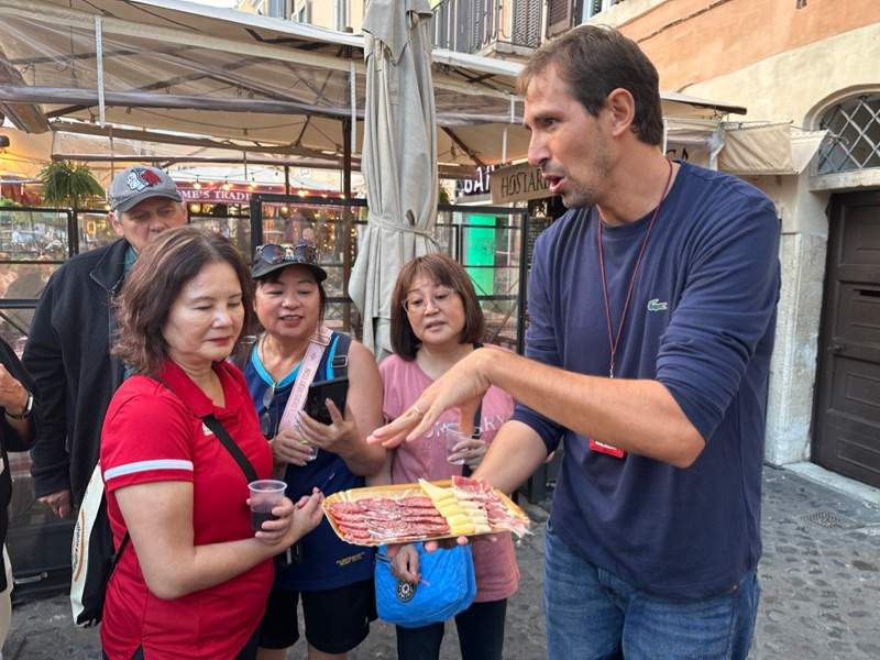 A group of tourists looking at fresh deli meats and cheeses while listening to their guide from The Tour Guy.