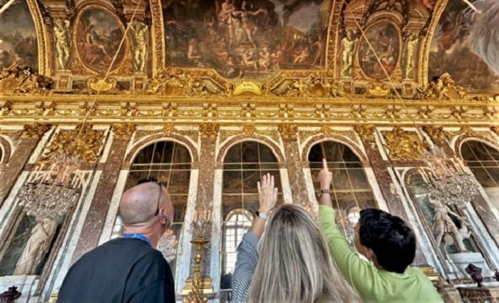 People pointing up to the ceiling of the palace of versailles in the hall of mirrors.