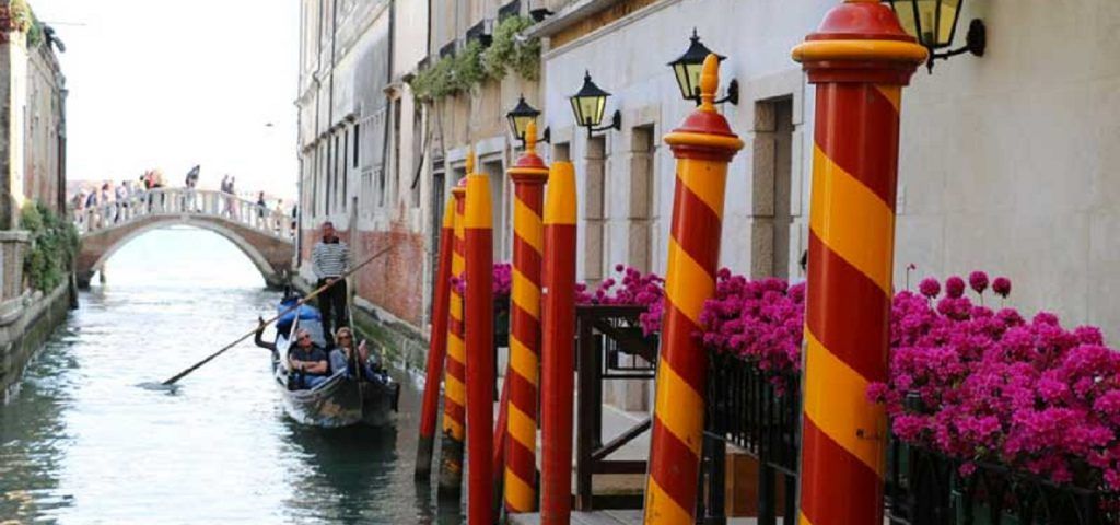 People on a gondola floating through the Venice canals. Red and Orange poles are in the foreground with some flowers.