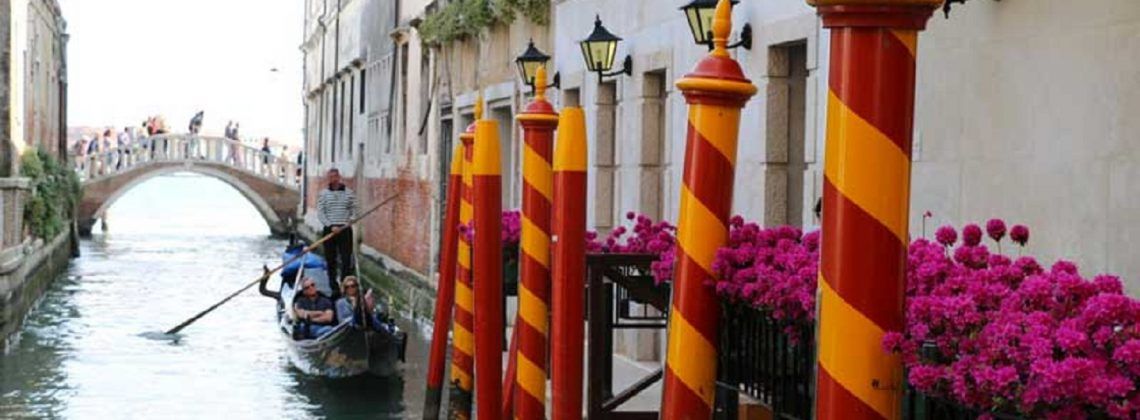 People on a gondola floating through the Venice canals. Red and Orange poles are in the foreground with some flowers.