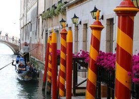 People on a gondola floating through the Venice canals. Red and Orange poles are in the foreground with some flowers.