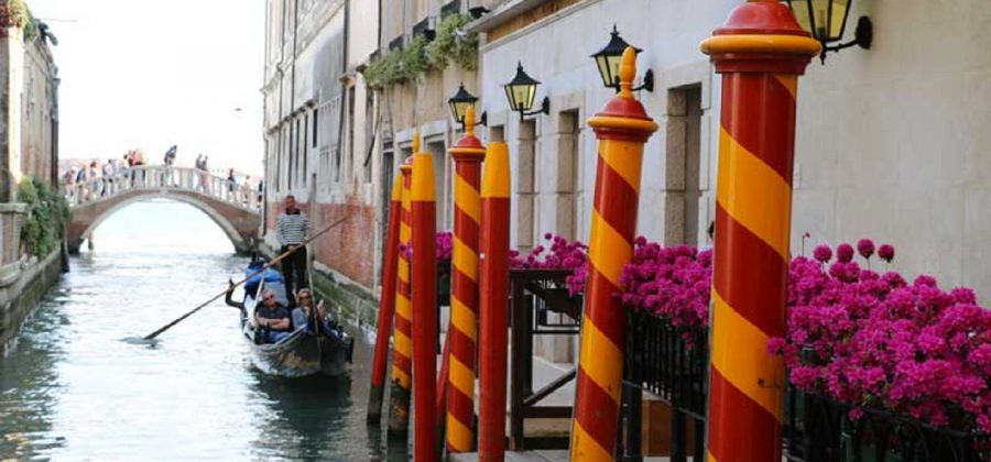 People on a gondola floating through the Venice canals. Red and Orange poles are in the foreground with some flowers.