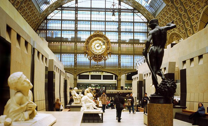 People in the central corridor of the Musee d'Orsay, Paris. In the background, there's the clock of the old railway station and, in the foreground, the David of Antonin Mercié seen from behind.