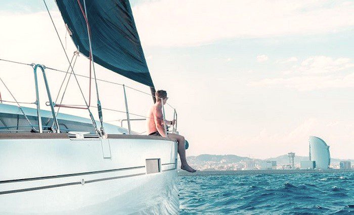 man sitting on the edge of a sailboat on a Barcelona boat cruise with views of the city in the background