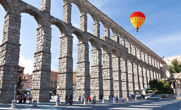 View of the Aqueduct of Segovia from the Plaza Azoguejo with people walking beneath the arches and a hot air balloon in the sky above the aqueduct.