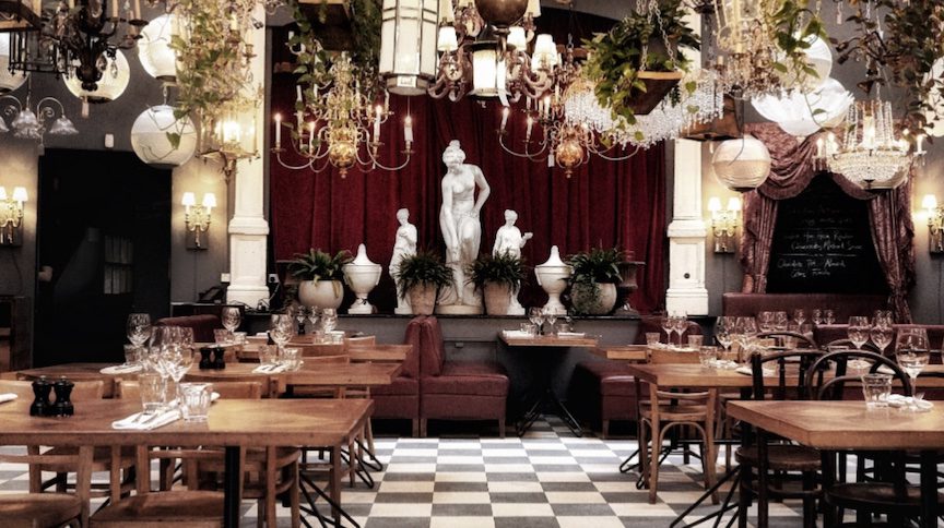 Chandeliers and antique statues surround wooden restaurant tables.