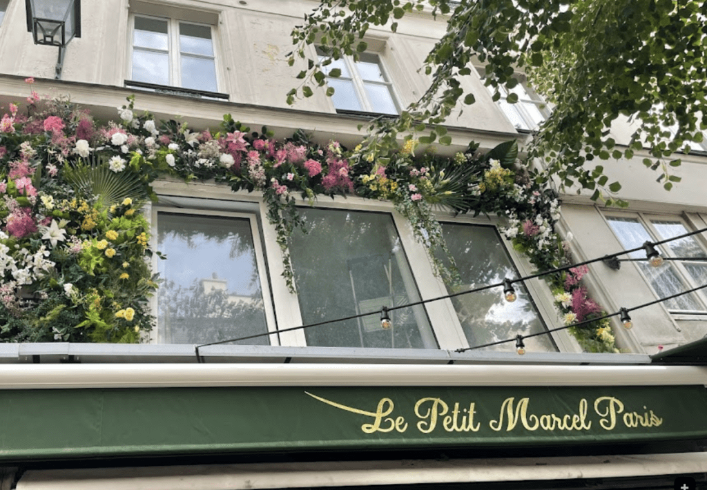 colorful flowers hang across a green sign that reads "Le Petit Marcel"