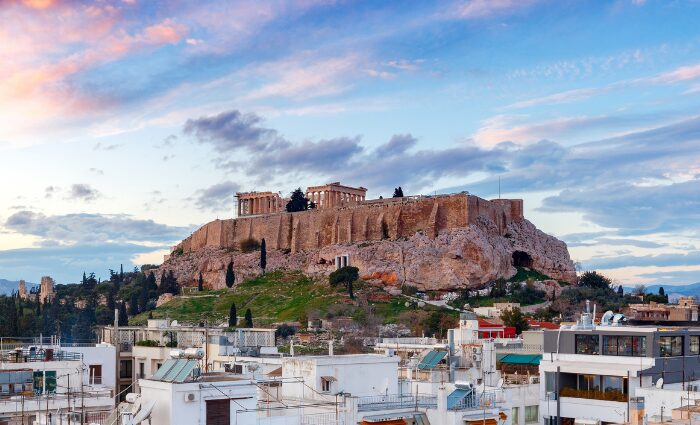 The Acropolis of Athens rising above the city rooftops at sunset, with the Parthenon visible at the top of the hill